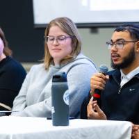four college student panelists sitting at the front of the students at the front of the conference room, one male student speaking while the others listen